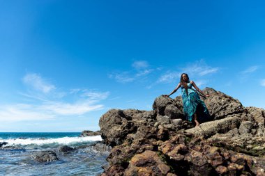 Mavi elbiseli ve örgülü saçlı güzel bir kadın kumsalın tepesinde oturmuş mavi gökyüzüne karşı güneşleniyor..