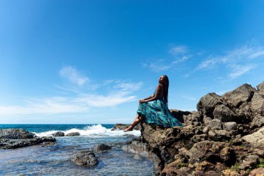 Mavi elbiseli ve örgülü saçlı güzel bir kadın kumsalın kenarında oturmuş mavi gökyüzüne karşı güneşleniyor..