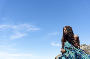 Mavi elbiseli, saçları örgülü, kumsalda kayalıklarda oturmuş mavi gökyüzüne bakan güzel bir kadının portresi..