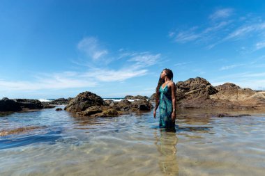 Mavi elbiseli ve örgülü saçlı güzel bir kadın sahilde suyun üzerinde duruyor mavi gökyüzüne karşı poz veriyor..
