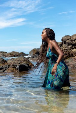 Mavi elbiseli ve örgülü saçlı güzel bir kadın sahilde suyun üzerinde duruyor mavi gökyüzüne karşı poz veriyor..