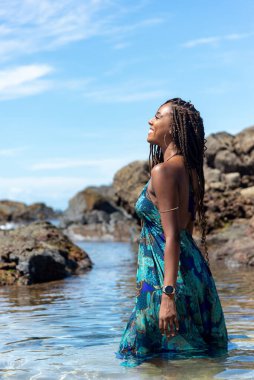 Mavi elbiseli ve örgülü saçlı güzel bir kadın sahilde suyun üzerinde duruyor mavi gökyüzüne karşı poz veriyor..