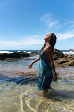 Mavi elbiseli ve örgülü saçlı güzel bir kadın sahilde suyun üzerinde duruyor mavi gökyüzüne karşı poz veriyor..