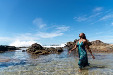 Mavi elbiseli, saçları örgülü, kumsalda duran, yürüyen ve mavi gökyüzüne bakan güzel bir kadın..