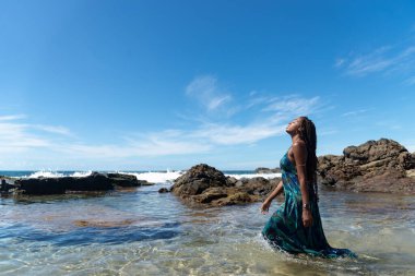 Mavi elbiseli, saçları örgülü, kumsalda duran, yürüyen ve mavi gökyüzüne bakan güzel bir kadın..