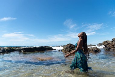Mavi elbiseli, saçları örgülü, kumsalda duran, yürüyen ve mavi gökyüzüne bakan güzel bir kadın..