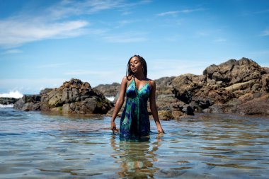 Mavi elbiseli güzel bir kadın ve kumsalda saçları örgülü bir kadın. Arka planda kaya ve mavi gökyüzüne karşı fotoğraf için poz veriyor..