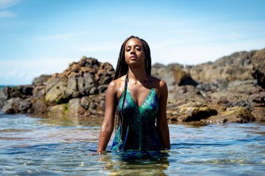 Sahil suyunda mavi elbiseli ve örgülü güzel bir kadın kameraya doğru yürüyor arkadaki kayalara ve mavi gökyüzüne doğru.