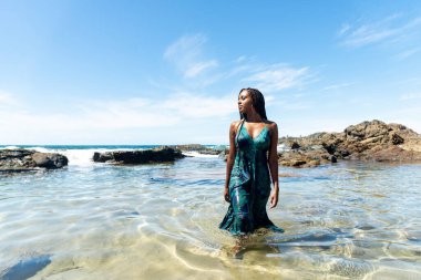 Sahil suyunda mavi elbiseli ve örgülü güzel bir kadın kameraya doğru yürüyor arkadaki kayalara ve mavi gökyüzüne doğru.