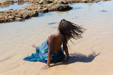 Mavi elbiseli güzel bir kadın kumsalda sırtı örgülü bir şekilde oturuyor. Seyahat eden kişi.