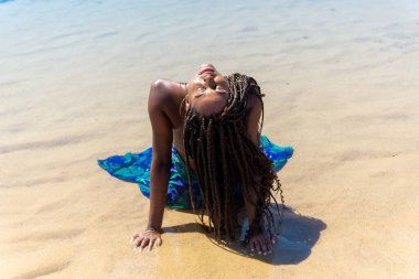 Mavi elbiseli, saçları örgülü, sırtı kapalı bir şekilde kumsalda oturan güzel bir kadın. Seyahat eden mutlu kişi.