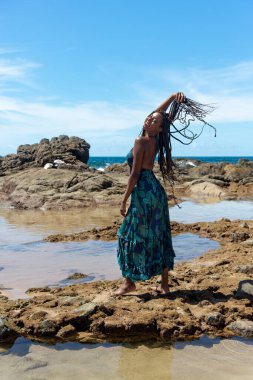 Güzel bir kadının portresi, kumsaldaki kayaların tepesinde mavi elbiseler giyip örgülü saçlarına dokunuyor. Seyahat eden mutlu kişi.