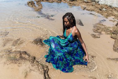 Saçları örgülü güzel bir kadın kumsalda uzun mavi bir elbiseyle güneşleniyor. Seyahat eden mutlu kişi.