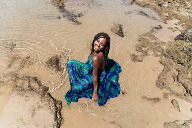 Saçları örgülü güzel bir kadın kumsalda uzun mavi bir elbiseyle güneşleniyor ve gülümsüyor. Seyahat eden mutlu kişi.