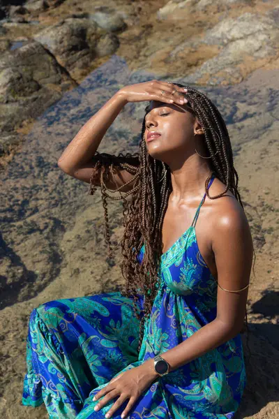 Mavi elbiseli güzel bir kadın kayalıklarda oturuyor, gözleri kapalı saçlarına dokunuyor, kumsalda güneşleniyor. Mavi gökyüzüne karşı.
