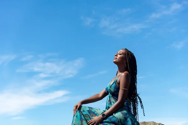 Beautiful woman in blue clothes and braided hair sitting sunbathing against blue sky.