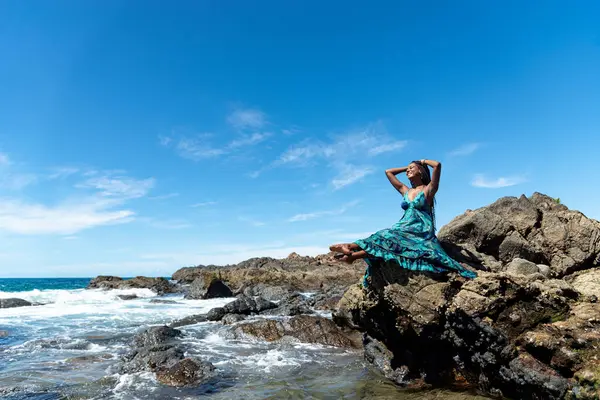 Beautiful woman in blue clothes sitting on the edge of the beach rocks touching her hair braids against blue sky.