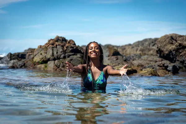 Woman in blue clothes and braided hair with her body in the beach water throwing water upwards against rocks and blue sky in the background.