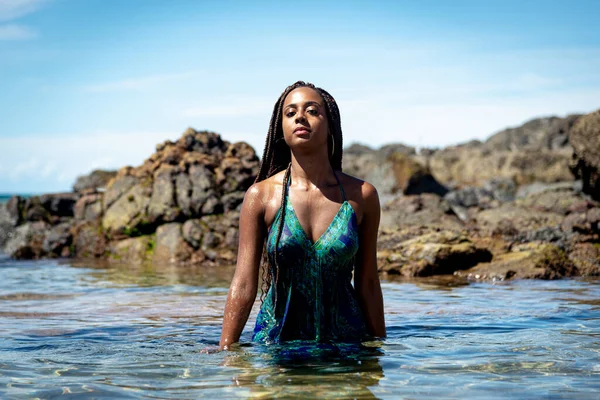 Sahil suyunda mavi elbiseli ve örgülü güzel bir kadın kameraya doğru yürüyor arkadaki kayalara ve mavi gökyüzüne doğru.