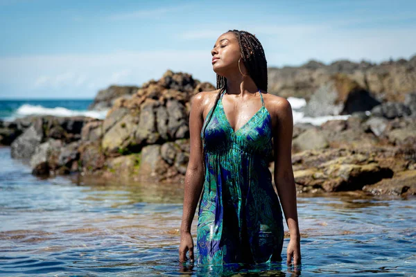 Sahil suyunda mavi elbiseli ve örgülü güzel bir kadın kameraya doğru yürüyor arkadaki kayalara ve mavi gökyüzüne doğru.