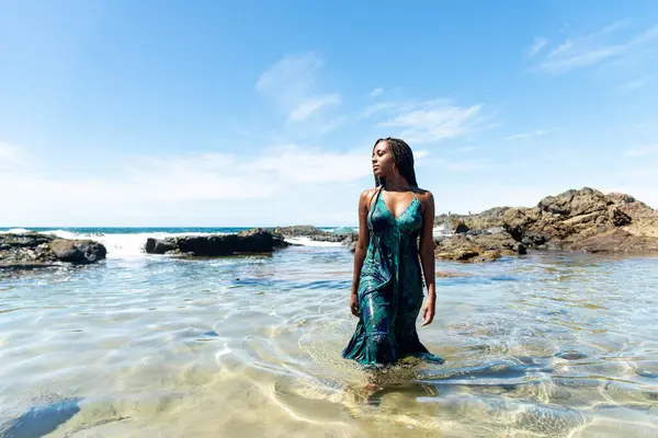 Sahil suyunda mavi elbiseli ve örgülü güzel bir kadın kameraya doğru yürüyor arkadaki kayalara ve mavi gökyüzüne doğru.