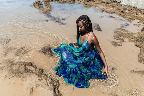 Saçları örgülü güzel bir kadın kumsalda uzun mavi bir elbiseyle güneşleniyor. Seyahat eden mutlu kişi.