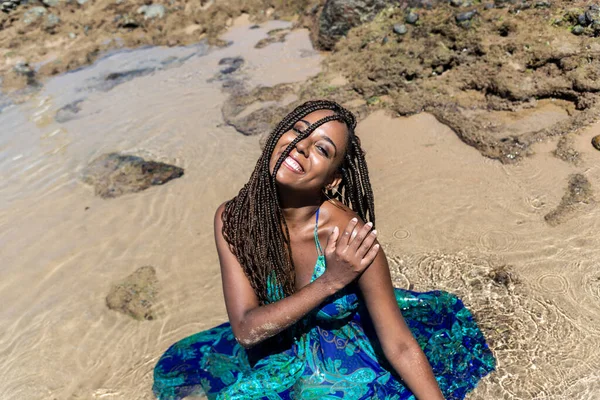 Güzel bir kadının portresi, mavi elbiseli ve örgülü, sahilde oturmuş fotoğraf için poz veriyor. Kumsalda güneşleniyor. Mavi gökyüzüne karşı.