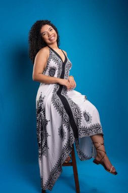Tahta bir taburede oturmuş fotoğraf için poz veren kıvırcık saçlı güzel bir genç kadın. Mavi arkaplanda izole.