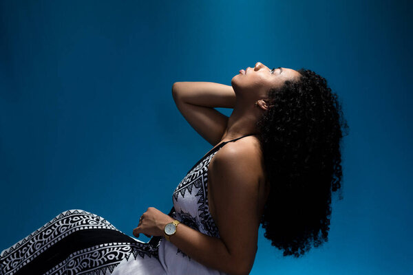 Portrait of beautiful woman, with curly hair, sitting on the floor, with head tilted back. Isolated on blue background.