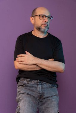 Sakallı, gözlüklü kel bir adamın portresi. Tahta taburede oturmuş fotoğraf için poz veriyor. Koyu leylak arka planda izole edilmiş.