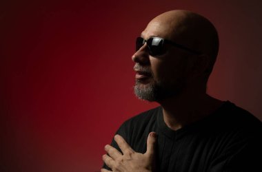 Güneş gözlüklü, yakışıklı, kel ve sakallı bir adam. Ciddi ve kendine güvenen bir poz veriyor. Kırmızı arkaplanda izole.