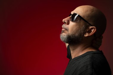 Yakışıklı, kel ve sakallı, güneş gözlüklü, ciddi ve kendine güvenen bir adam. Kırmızı arkaplanda izole.