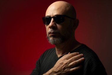 Güneş gözlüklü, yakışıklı, kel ve sakallı bir adam. Ciddi ve kendine güvenen bir poz veriyor. Kırmızı arkaplanda izole.