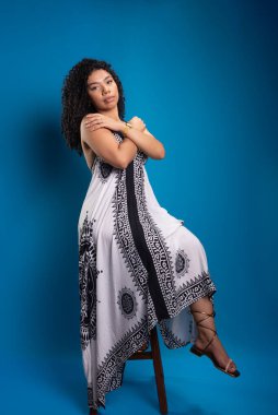 Tahta bir taburede oturmuş fotoğraf için poz veren kıvırcık saçlı güzel bir genç kadın. Mavi arkaplanda izole.