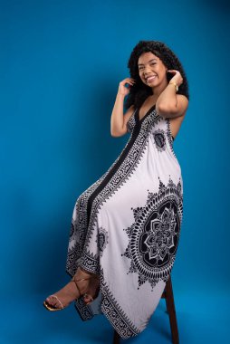 Tahta bir taburede oturmuş fotoğraf için poz veren kıvırcık saçlı güzel bir genç kadın. Mavi arkaplanda izole.