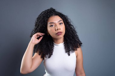 Genç ve güzel bir kadın kıvırcık saçlarına dokunuyor ve gri arka planda kameraya bakıyor..