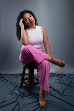 Kıvırcık saçlı, resmi giyinmiş güzel bir kadın tahta taburede oturmuş fotoğraf için poz veriyor. Gri arkaplanda izole.