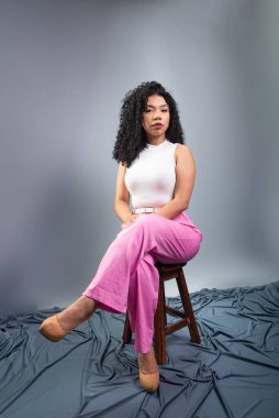 Kıvırcık saçlı, resmi giyinmiş güzel bir kadın tahta taburede oturmuş fotoğraf için poz veriyor. Gri arkaplanda izole.