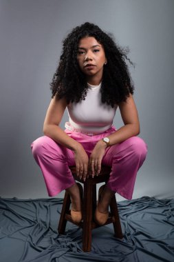 Güzel bir kadın, kıvırcık saçlı, gri arka planda oturmuş kameraya ciddi ciddi bakıyor..