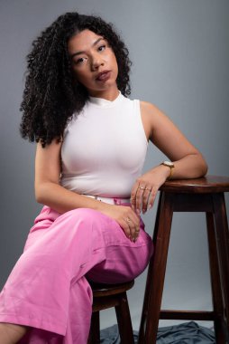 Güzel bir kadın, kıvırcık saçlı, gri arka planda oturmuş kameraya ciddi ciddi bakıyor..