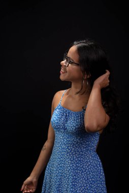 Gözlüklü güzel esmer kadın saçlarına dokunup poz veriyor. Siyah arka planda mavi elbiseler giyiyor..