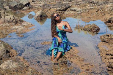 Güzel bir kadın, saç örgüleri ve mavi elbisesiyle kumsalda güneşlenen kayalarda oturuyor. Seyahat eden kişi.