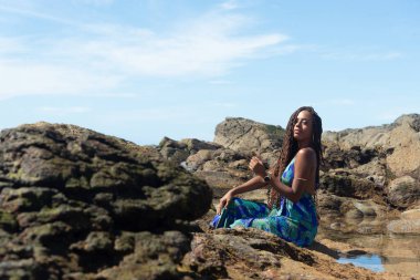 Güzel bir kadının portresi, örgülü ve mavi elbiseli, kumsalın kayalıklarında mavi gökyüzüne karşı poz veriyor..