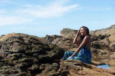 Güzel bir kadın, mavi elbiseli, elleri örgülü kayalıklarda oturuyor, kumsalda güneşleniyor ve yan tarafa bakıyor. Mavi gökyüzüne karşı.