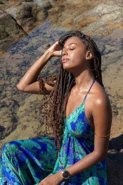 Mavi elbiseli güzel bir kadın kayalıklarda oturuyor, gözleri kapalı saçlarına dokunuyor, kumsalda güneşleniyor. Mavi gökyüzüne karşı.