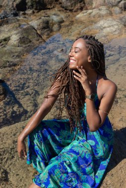 Güzel bir kadının portresi, örgülü ve mavi elbiseli, kumsalın kayalıklarında mavi gökyüzüne karşı poz veriyor..