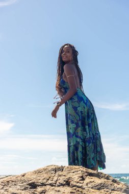 Güzel bir kadın, saç örgüleri ve mavi elbiseleriyle sahildeki kayalıklarda dikilip mavi gökyüzüne karşı poz veriyor..