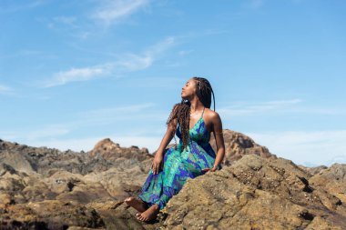 Güzel bir kadın, saç örgüleri ve mavi elbiseleriyle kumsalda güneşlenen kayalarda oturuyor ve mavi gökyüzüne karşı poz veriyor..