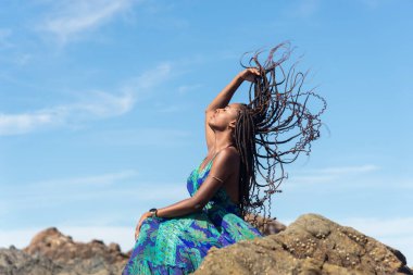Mavi elbiseli güzel bir kadın kumsalda kayalıklarda güneşleniyor ve mavi gökyüzüne karşı örgülerine dokunuyor..