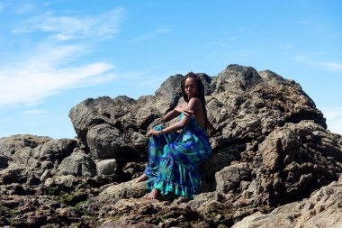 Mavi elbiseli, örgülü saçlı, kumsalın karanlık kayalarında oturmuş mavi gökyüzüne karşı güneşlenen güzel bir kadın..